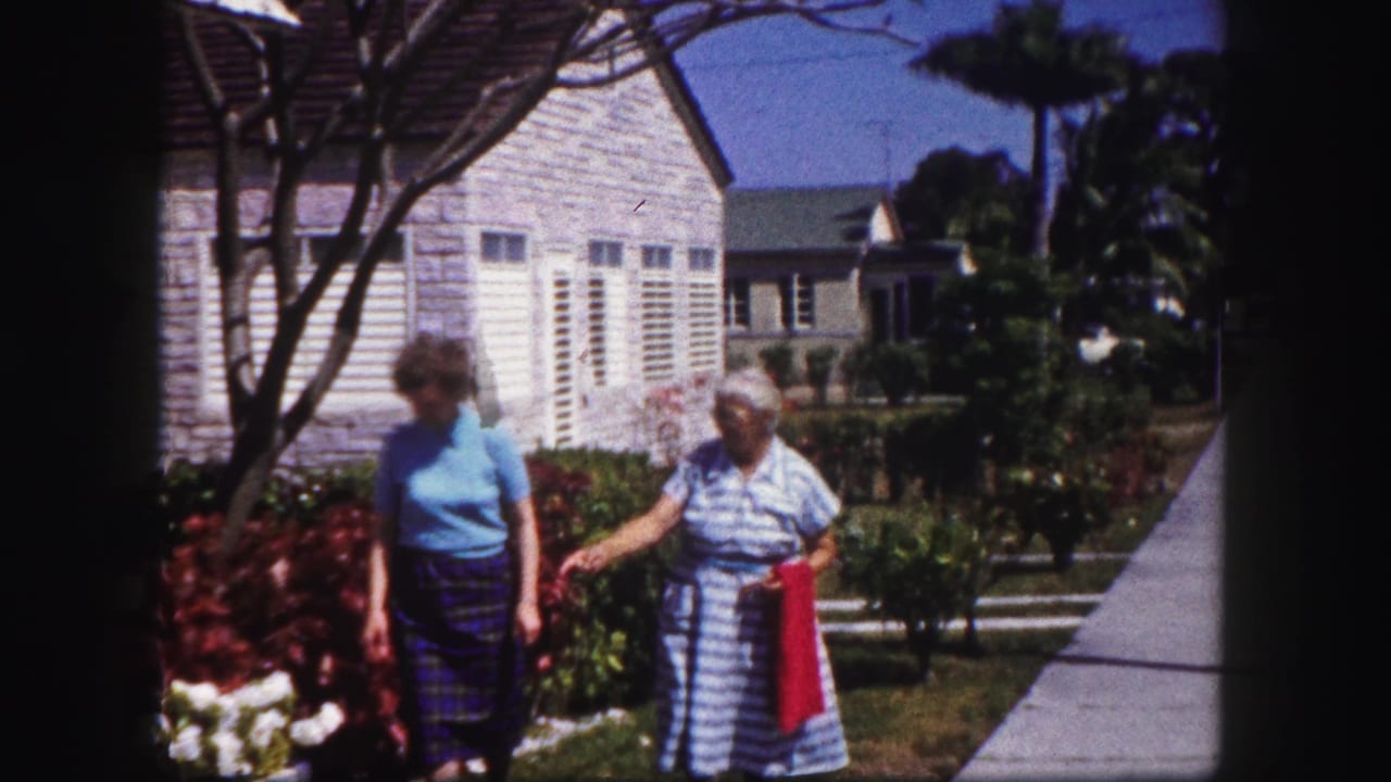 Two women walk beside flowers and a stone-faced house in a sunny residential yard.
