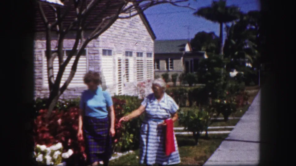 In Tampa, Florida, circa 1960, two adult women walk near flowering shrubs, a sidewalk, and a stone-faced house with white shutters.