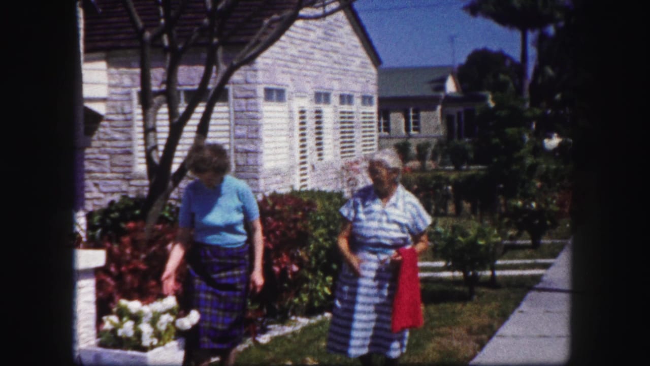 In the 1960 Tampa home-movie scene, two women pause near a planted border, lawn, sidewalk, and white-shuttered residential house.