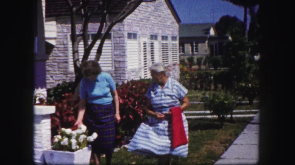 Two adult women in a Tampa residential garden scene look toward flowers near a concrete sidewalk and white-shuttered house, circa 1960.