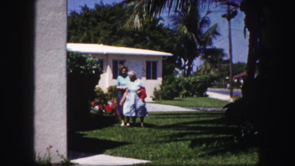 In a 1960 Tampa suburban yard, two women walk together across grass near palms, shrubs, a sidewalk, and a low light-colored house.