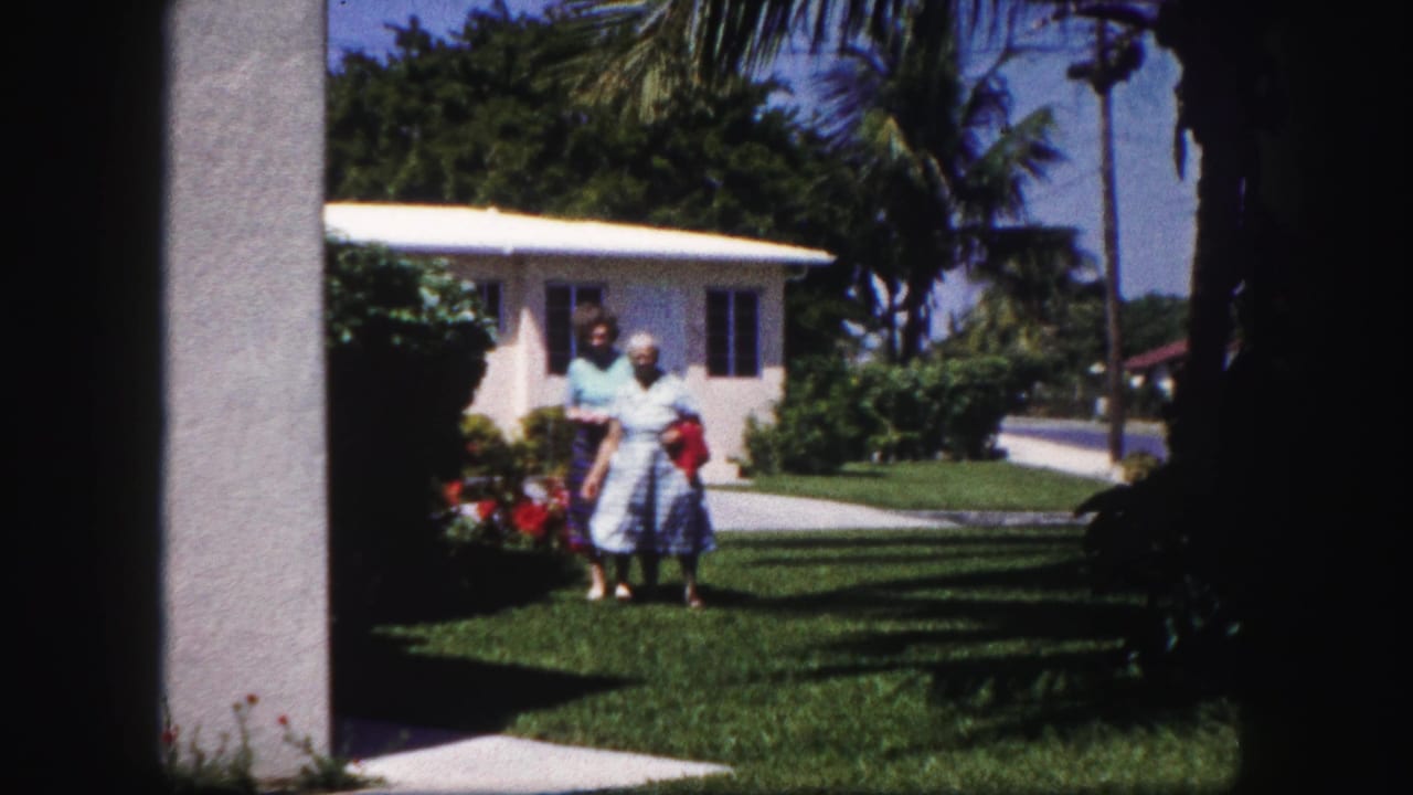 Two women in 1960 Tampa stand on a sunny suburban lawn near a white stucco house and palm trees, seen through dark doorway framing.
