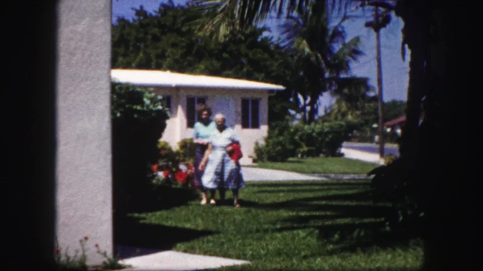 Two women in 1960 Tampa stand on a sunny suburban lawn near a white stucco house and palm trees, seen through dark doorway framing.