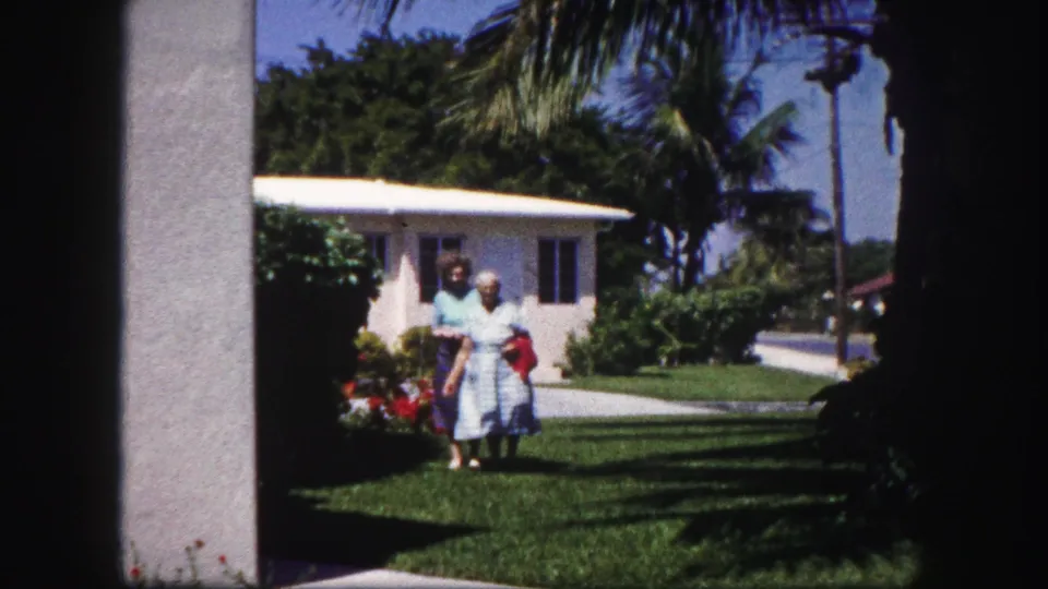 Two women in a sunny Tampa residential yard stand near a white stucco house, with palms, shrubs, and a sidewalk visible around them.