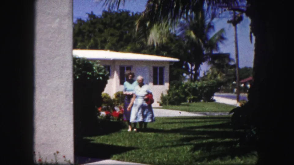 Two women in period clothing walk across a sunny Tampa lawn near a white stucco house, framed by shadowed edges of a doorway.