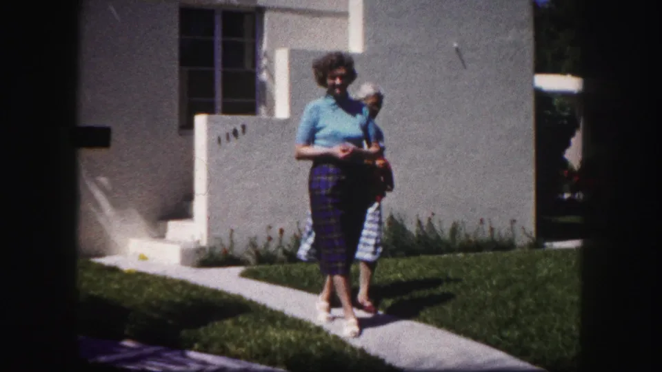 A woman in a blue top and plaid pants walks toward the camera outside a Tampa stucco house, with another woman partly visible behind her.