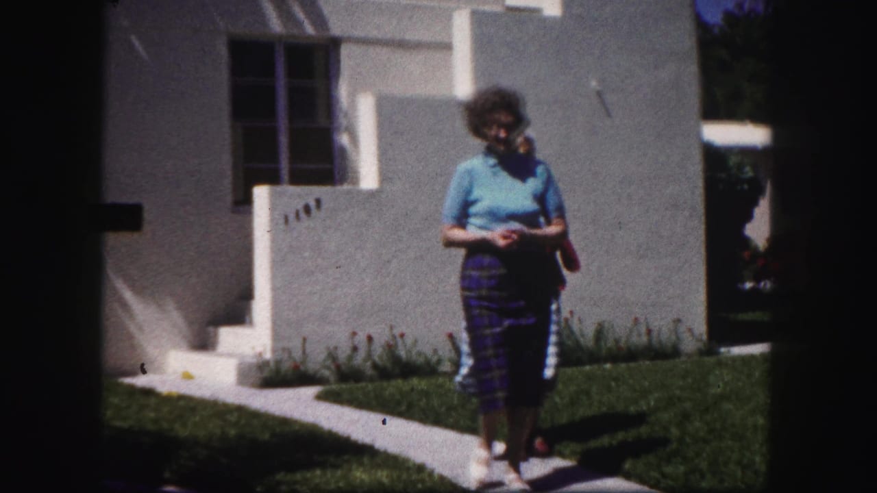 Older woman walking on a sidewalk beside a white stucco home and lawn.