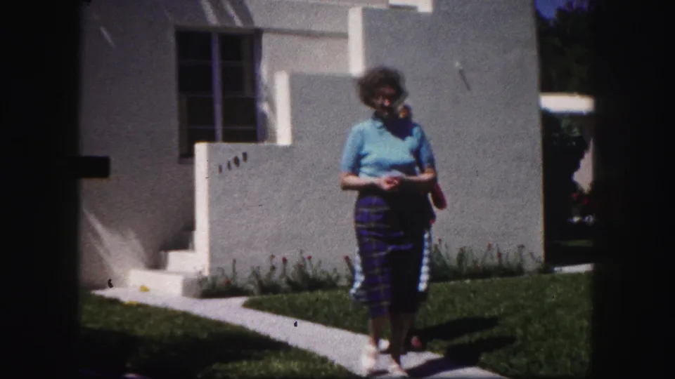 In the supplied 1960 Tampa footage, an older woman in a blue top and plaid skirt walks near a white stucco residence.