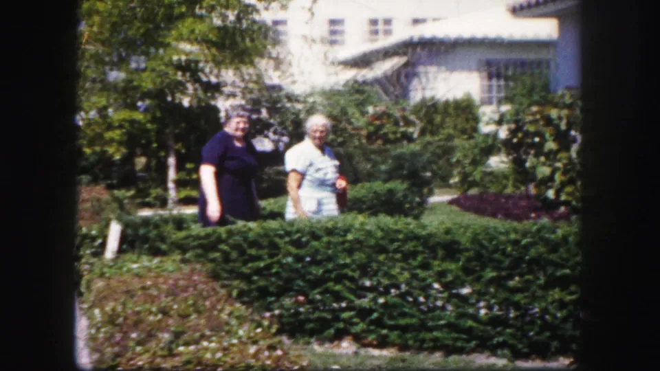 Two older women face the camera from behind a hedge in a sunlit Tampa residential garden, with stucco homes in the background.