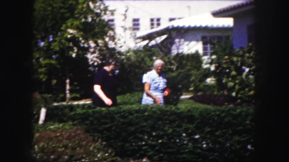 Two women appear near a front-yard hedge and flowering shrubs in 1960 Tampa 8mm footage, one holding a red cloth.