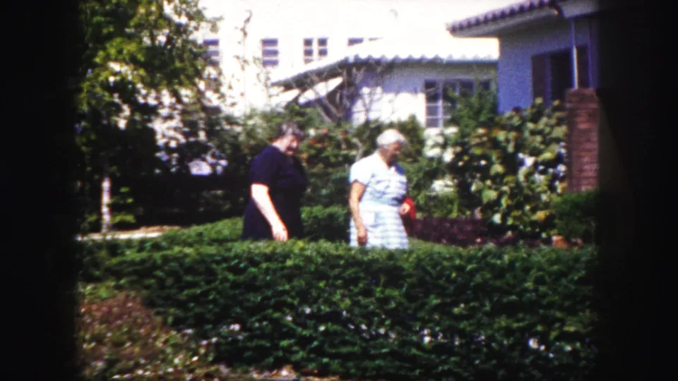Two women are seen beside a manicured hedge and garden plantings outside a Tampa house in grainy 1960 8mm footage.