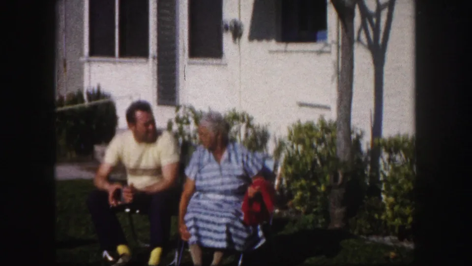 A man and elderly woman sit together on the lawn beside a Tampa residence, with shrubs, windows, and a tree behind them.