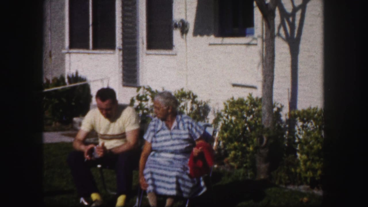 Man and woman seated on lawn chairs outside a white house in sunlight.