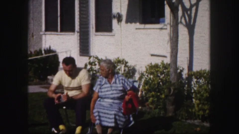 In supported 1960 Tampa home-movie footage, a man in a yellow striped shirt and a woman in a striped dress sit outside near shrubs.