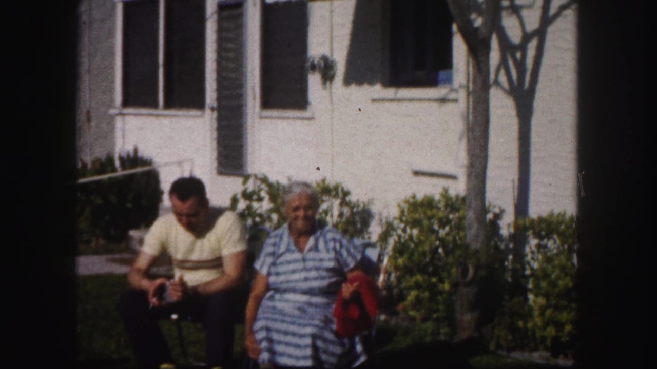 The man looks downward while the woman sits beside him in a striped dress outside a white house in Tampa, Florida.