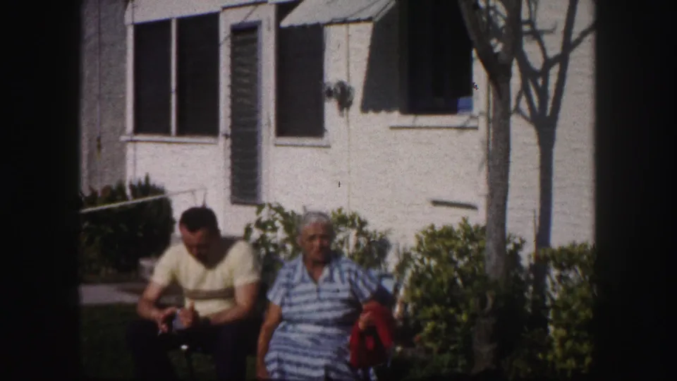 A 1960 home-movie frame shows two seated adults outdoors, with the woman facing forward and the man leaning slightly down.