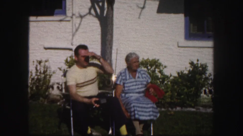 In the final frame, the man raises a hand to his sunglasses while the woman sits nearby holding or resting beside a red cloth.
