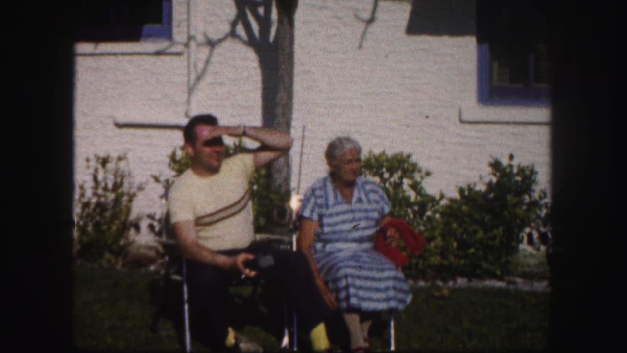 In 1960 Tampa home-movie footage, a man shields his eyes while seated beside a woman in a striped dress on a sunny lawn.