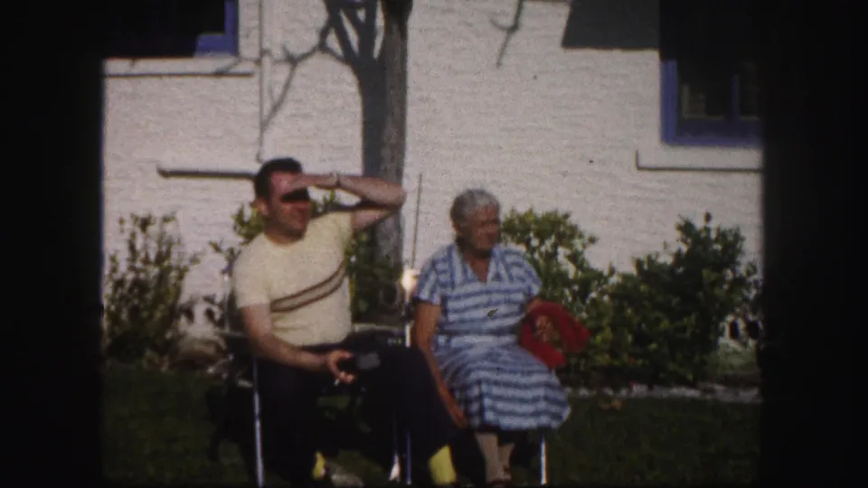 In 1960 Tampa home-movie footage, a man shields his eyes while seated beside a woman in a striped dress on a sunny lawn.