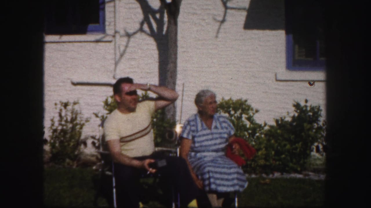 A man in a yellow striped shirt and a woman in a blue striped dress sit in lawn chairs outside a Tampa house in 1960 footage.