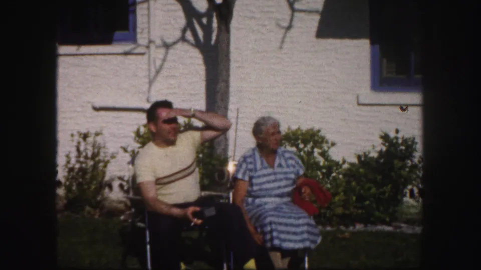 A man in a yellow striped shirt and a woman in a blue striped dress sit in lawn chairs outside a Tampa house in 1960 footage.