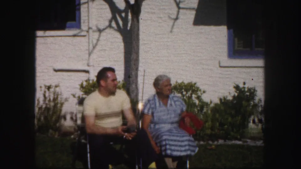 The seated couple appears to converse on a green lawn, with a white stucco house and shrubs behind them in 1960 Tampa footage.
