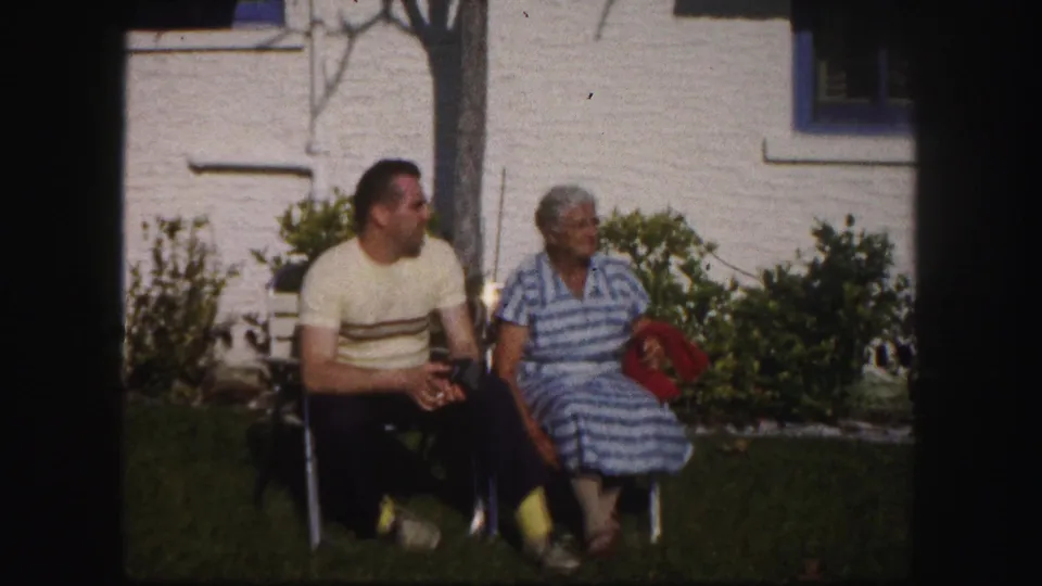 A man and woman sit close together in lawn chairs outside a blue-trimmed white house, captured in 1960 Tampa home-movie footage.