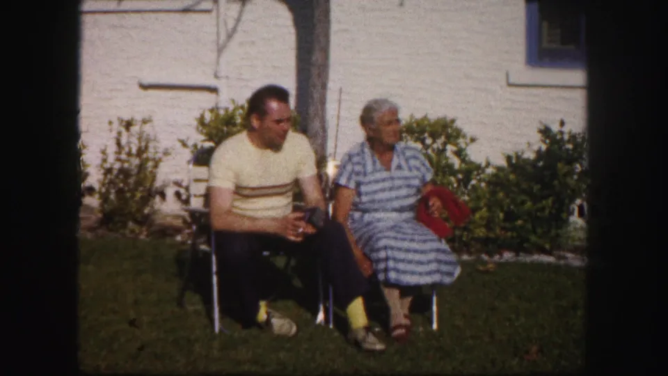 In 1960 Tampa home-movie footage, a man and older woman sit outdoors on folding chairs near a light-colored building and shrubs.