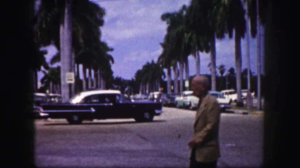 A black mid-century sedan passes through a palm-lined Tampa street while an older man walks near the foreground.