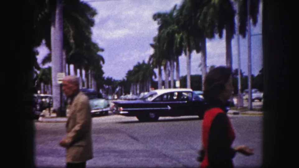 The street view continues with a dark sedan, parked cars, tall palms, and pedestrians moving through the foreground.