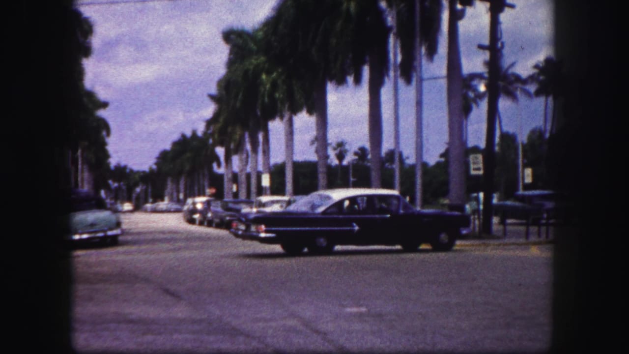 The sedan continues across the 1960 Tampa intersection, framed by parked cars, palms, and dark 8mm gate edges.