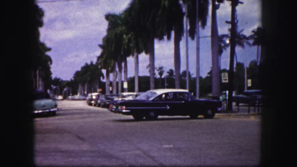 The sedan continues across the 1960 Tampa intersection, framed by parked cars, palms, and dark 8mm gate edges.