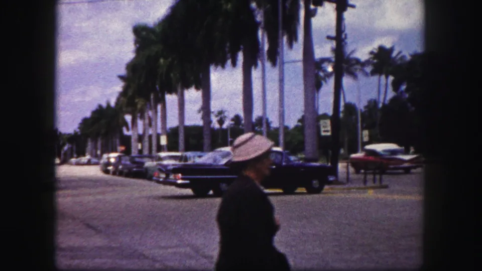 A pedestrian wearing a light hat crosses the foreground as the 1960 Tampa street scene continues behind them.