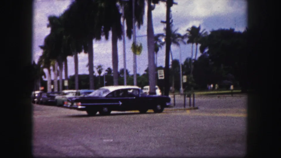 The sedan appears farther through the 1960 Tampa intersection, with palm trunks, parked cars, and curbside signs visible.