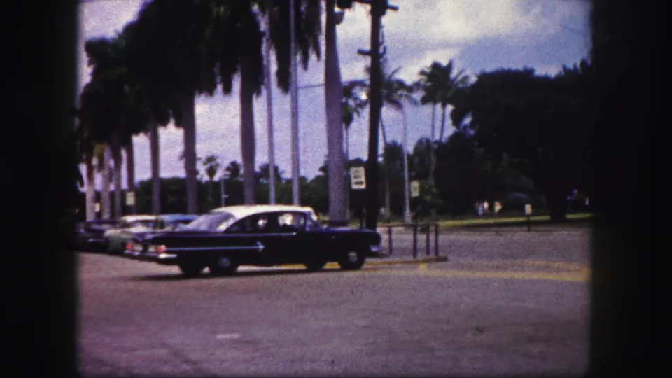 1960 Tampa street view with a dark sedan, parked cars, palm trees, traffic signs, and a paved intersection.