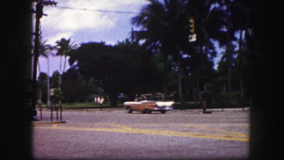 A light-colored convertible passes across the Tampa intersection, with palm trees, road markings, and distant people in the background.