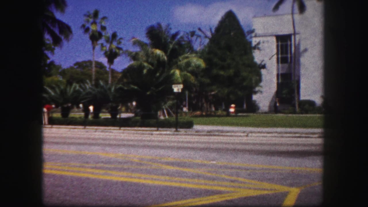 1960 Tampa street view with yellow road markings, palm landscaping, and a large white building seen from an 8mm home-movie frame.