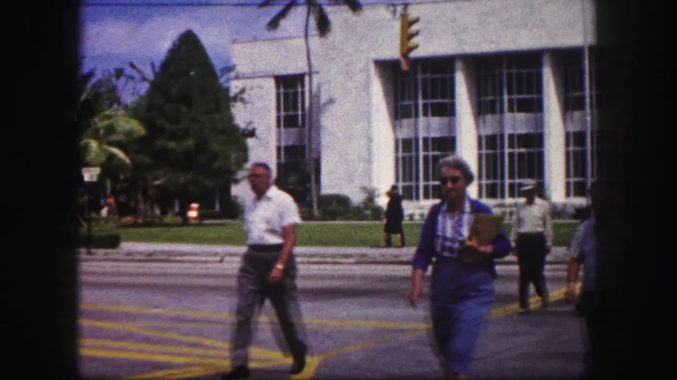 Pedestrians, including a woman carrying a folder, cross a Tampa street before a white building with tall windows and palm landscaping.