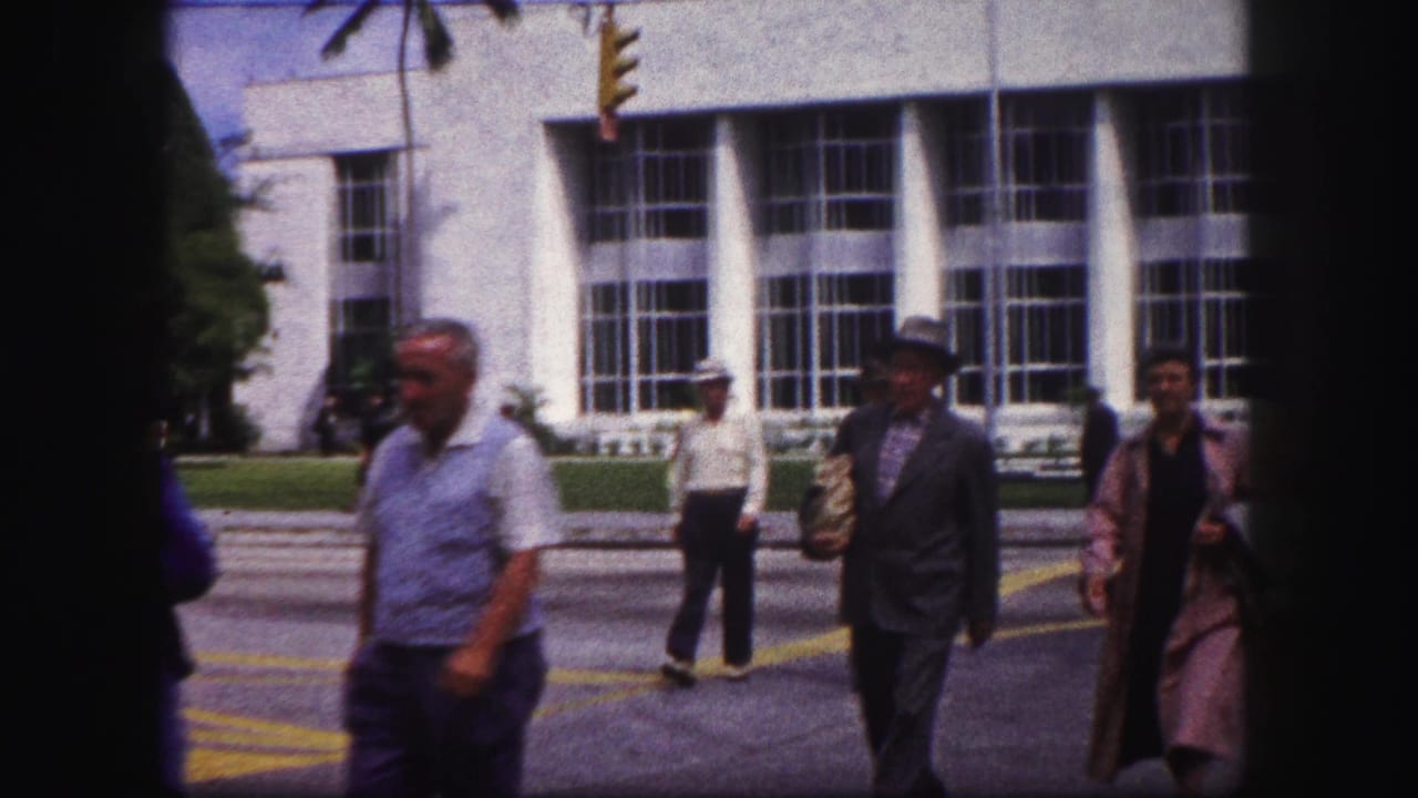 Pedestrians cross a sunlit street in front of a large-window modernist building.