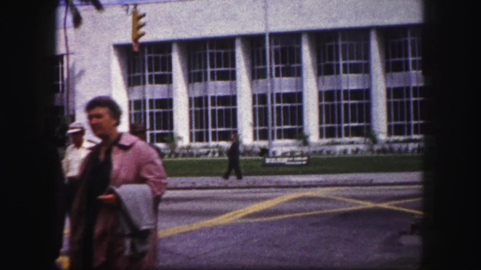 1960 Tampa street frame showing a woman passing close to camera, with yellow road markings and a modernist facade behind her.