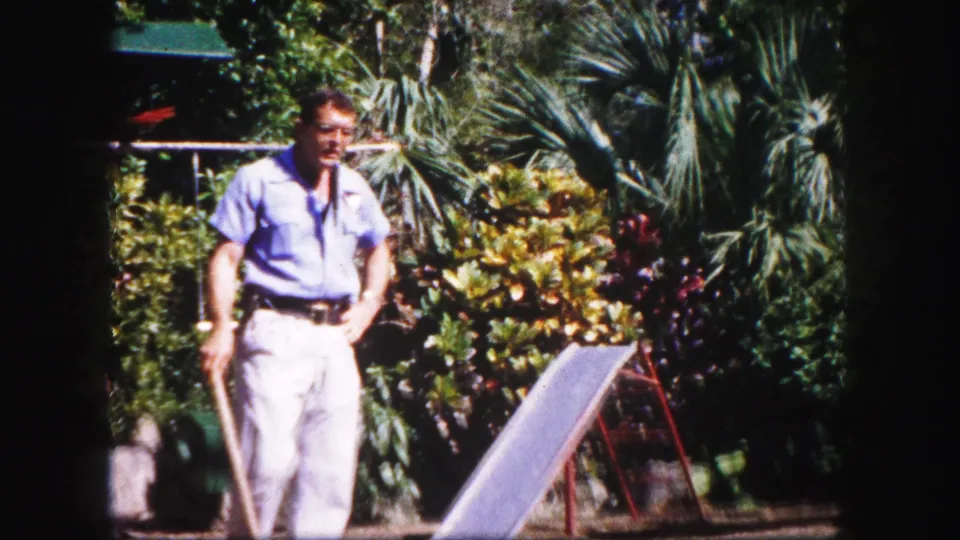 1960 Tampa frame of the man standing in a tropical garden, the macaw no longer prominent, with palms and a slide-like structure nearby.