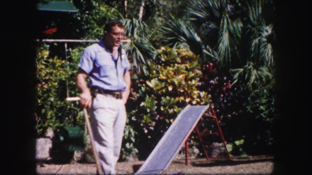 Man in a garden holds a long tool near a small ramp and tropical plants.