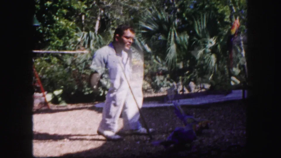 An adult man in white trousers moves across the tropical garden, holding a long-handled tool or lead in this 1960 Tampa footage.