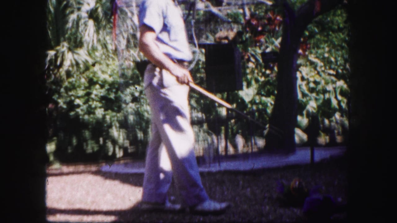 Man in light clothing walks through a tropical garden holding a long object near a birdcage.