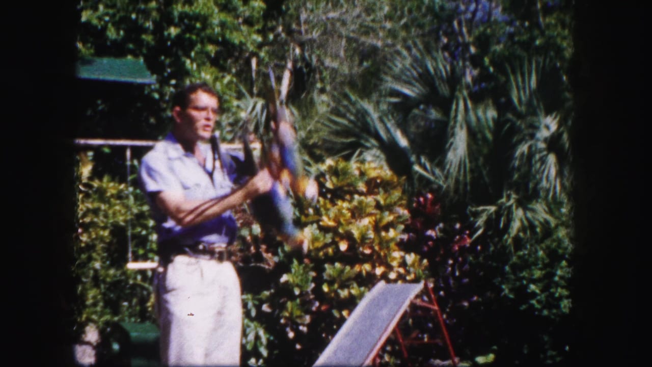 Man in a light shirt holds a colorful parrot in a tropical garden.