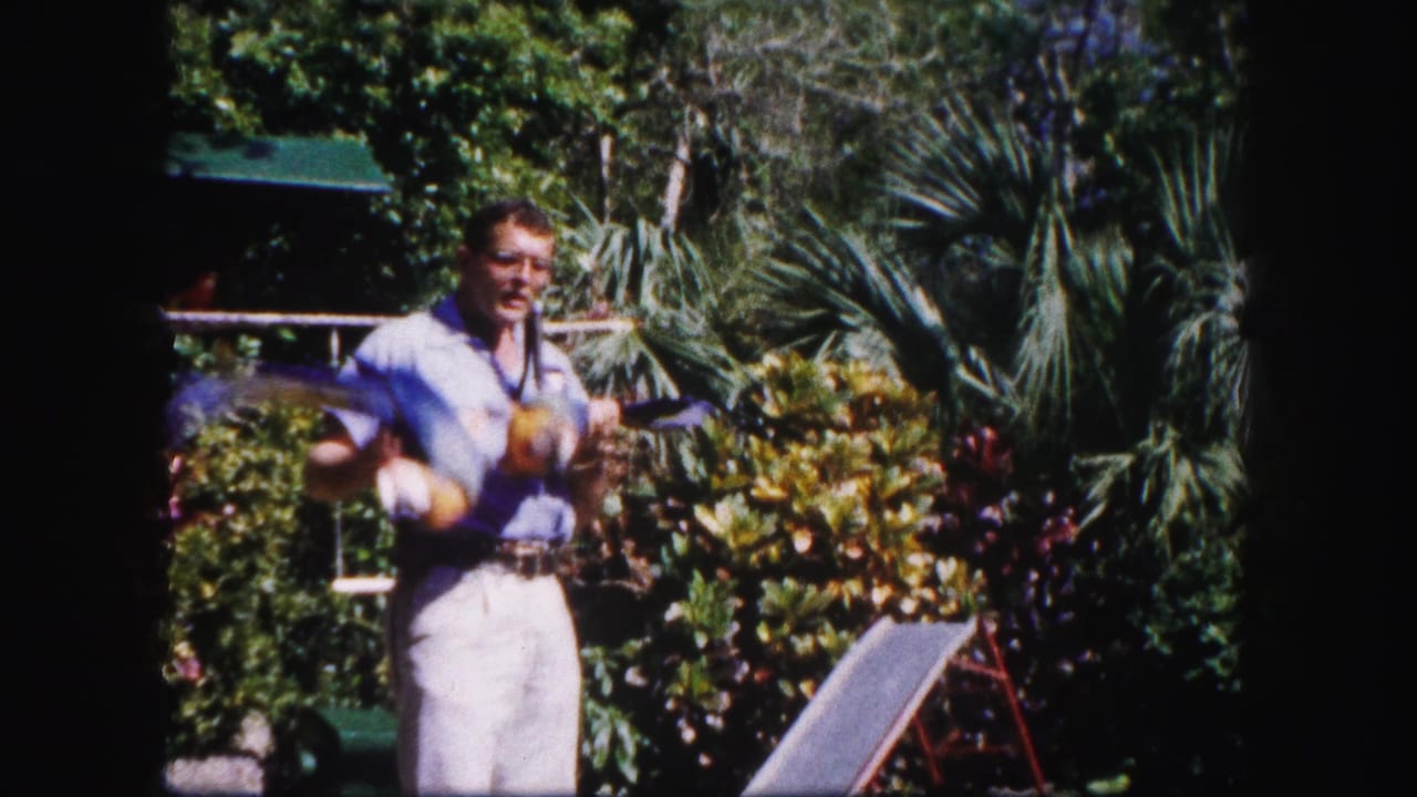 In 1960 Tampa, an adult man in light clothing walks through lush garden foliage with a parrot perched on a stick.