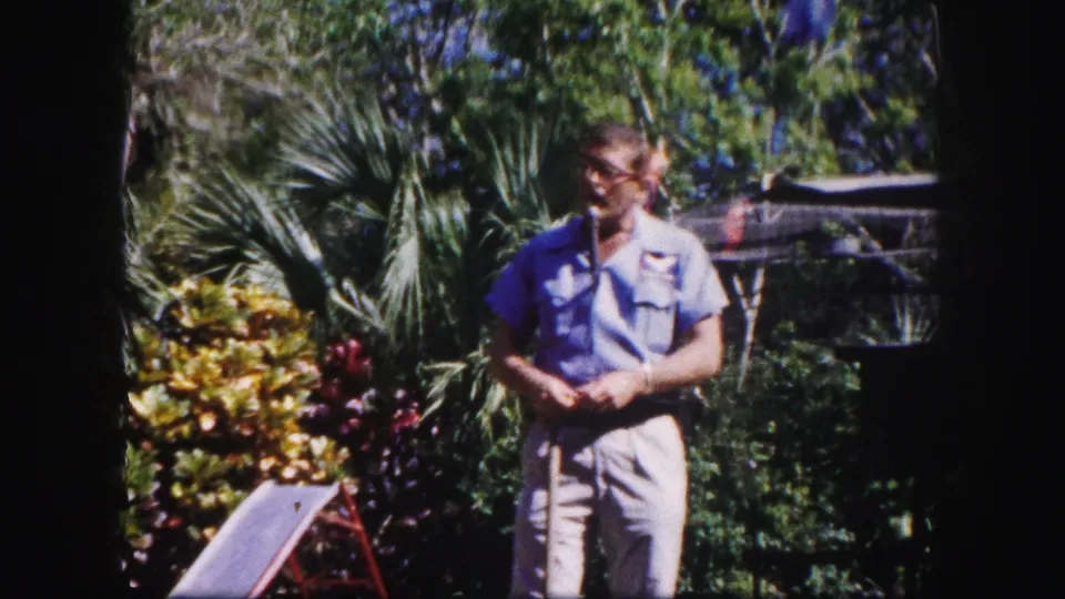 The zookeeper stands with a stick before tropical planting and aviary wire, with the bird mostly out of frame.