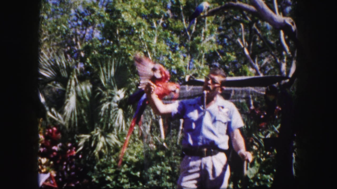 Man in glasses holds a red macaw on his arm in a leafy enclosure.