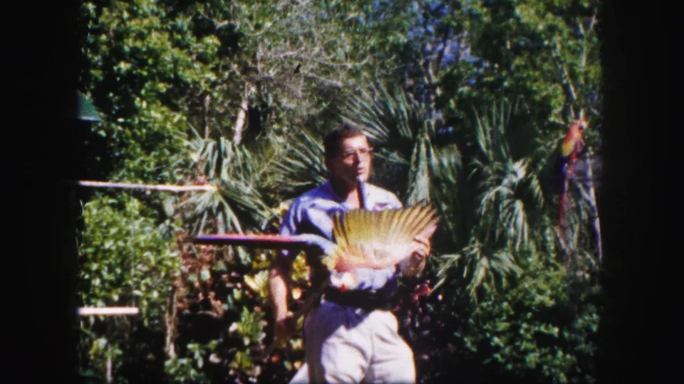 A 1960 Tampa home-movie frame shows a guide presenting a parrot with open wings in a lush outdoor garden.
