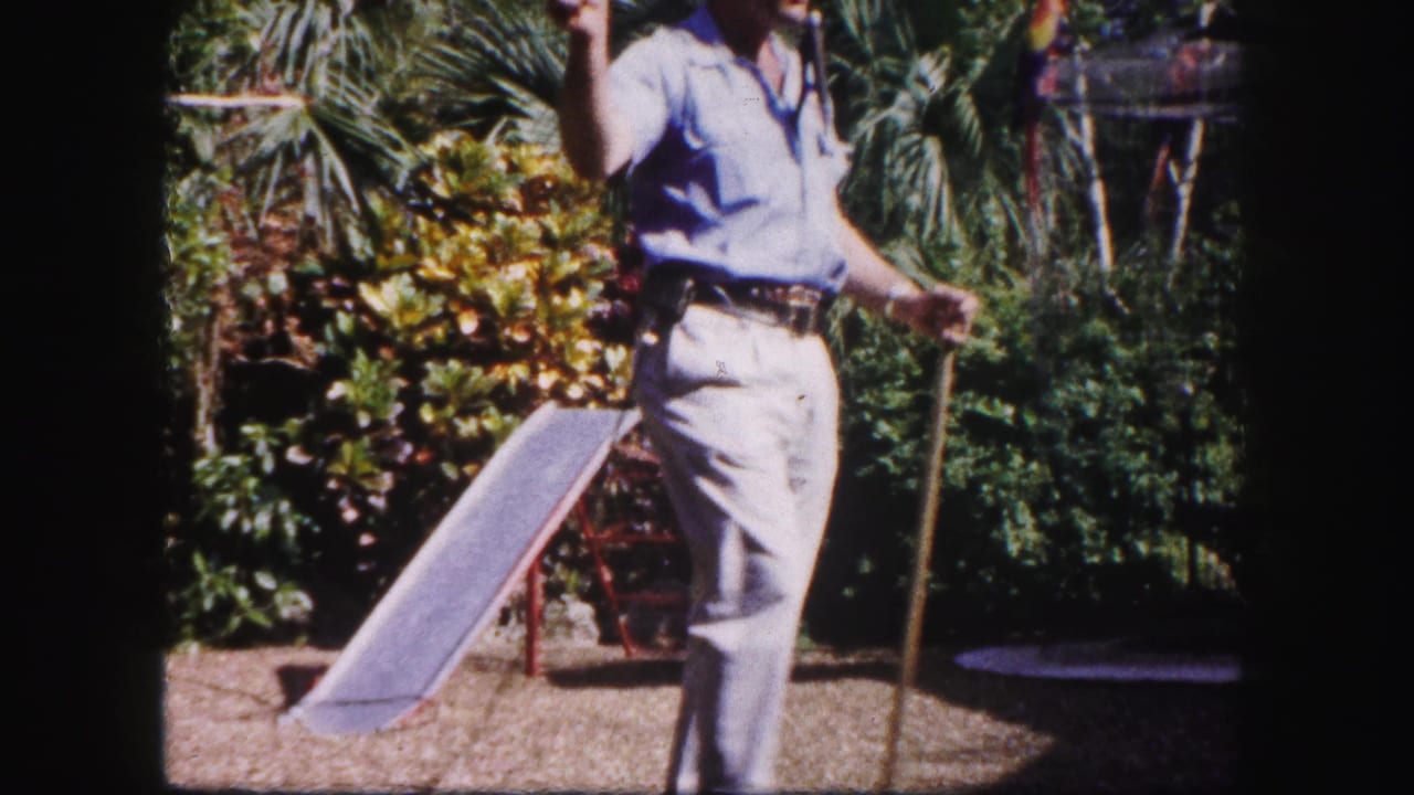 In 1960 Tampa zoo footage, a man in a blue shirt and khaki pants stands in a planted enclosure area with a pole and colorful parrot nearby.